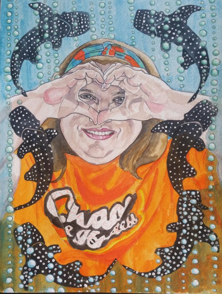 ID: Woman framing her eyes with her hands in a heart shape. She is smiling. She has shoulder length hair and is wearing a headband in orange, blue and turquoise. Her T shirt is orange and has “Chaos and Kindness” written on it, in white with a black surround. She is framed by bubbly sea water which runs across the bottom of her t shirt creating a greeny colour and is also behind her head which is blue. There are bubbles coming up in columns which are greeny/blue in colour on the top half of the paper and blue on the bottom half. She is framed by whale sharks dancing round her.