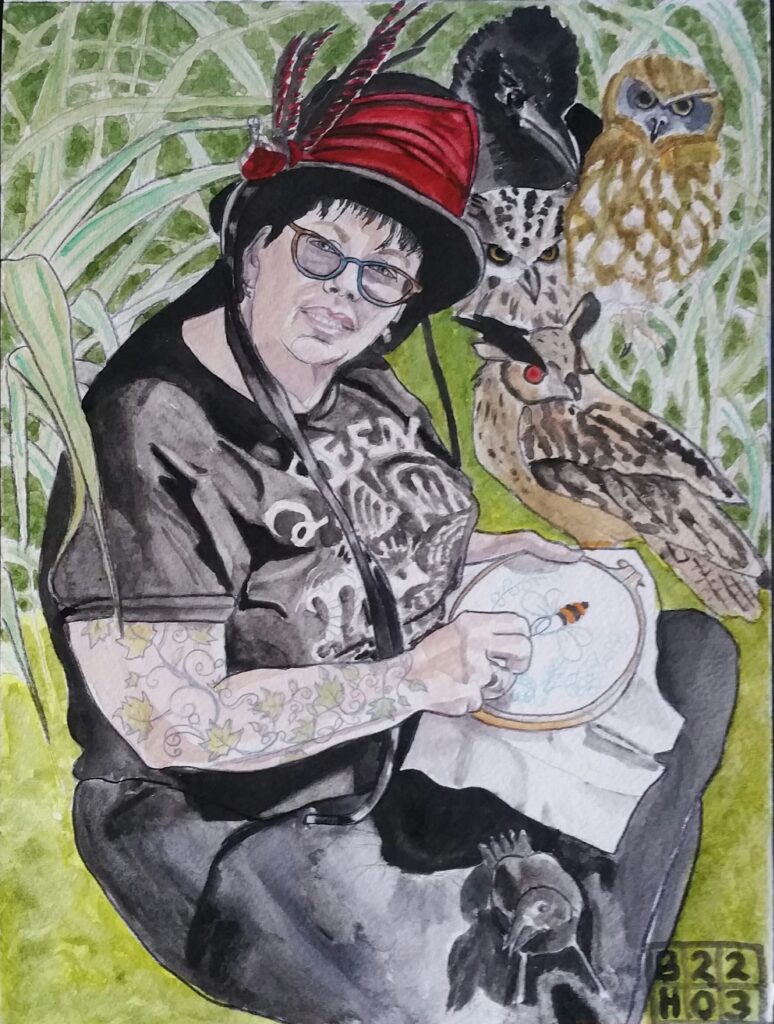 ID Woman sat cross legged on the grass, wearing a black top hat decorated with red fabric and feathers in red and black. She is wearing glasses and a black top with white writing and patterns on it and black trousers. she is turned to one side but has her head turned to look at the viewer. Down her near arm is a tattoo of lnter twirling leaves and tendrils. She is holding an embroidery ring with fabric stretched across it and is sewing a bee. She has a crow sat on one knee. In the background are long leaves and to the right of her can be seen a collage of owls and a crow.