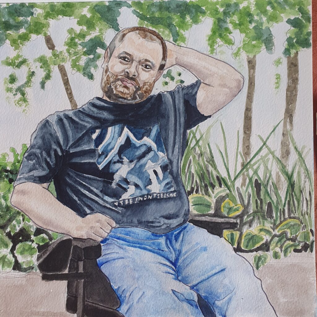 portrait of round headed man with short hair and a short beard and moustache - he has one hand behind his head and the other one is resting in his lap. He is wearing a t-shirt and jeans and is sat in a wheelchair, in the background can be seen general foliage.