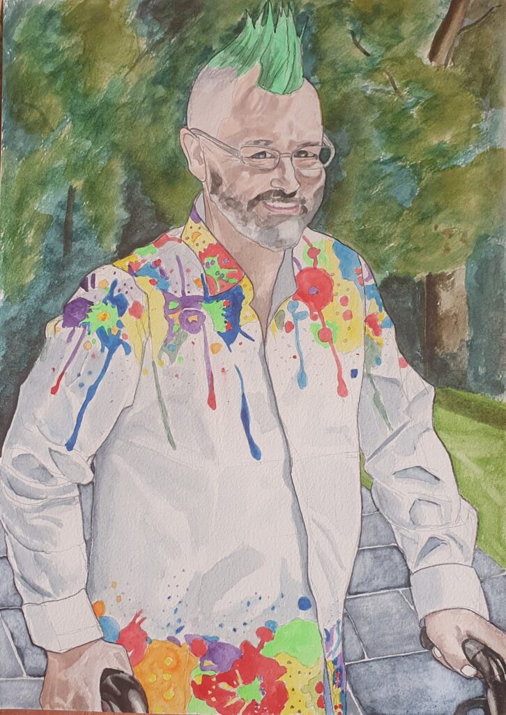 ID Man walking though park with a walker, He has a green mohican, glasses and an engaging smile, and is wearing a white long sleeved shirt with bright coloured patterns and runs of colour, at the top and bottom of the shirt. In the upper background are trees and the lower background is dark grey flagstones .