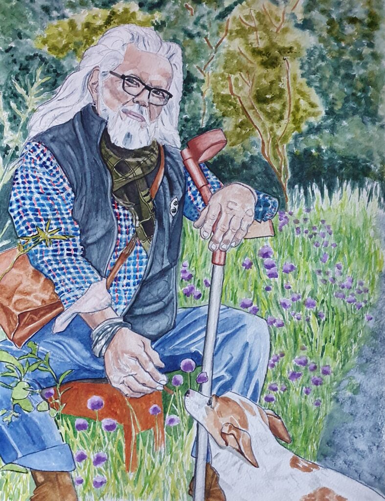 ID Man sat down outside on a stool - he has long hair, glasses and a beard, he is wearing a shirt neckerchief and woollen waistcoat and is holding a treat out for his dog which is a Jack Russell. His arm is leaning on the handle of his crutch. In the background is foliage, leaves and flowers