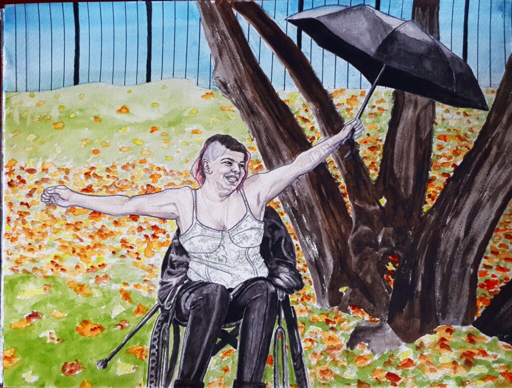 ID Non-binary person sat in wheelchair, they have a mohican hairstyle and they are flinging their arms out, black brolly in one hand, in an expression of joy. Behind them are trees and a leafy covered ground. And even further in the distance is iron railings