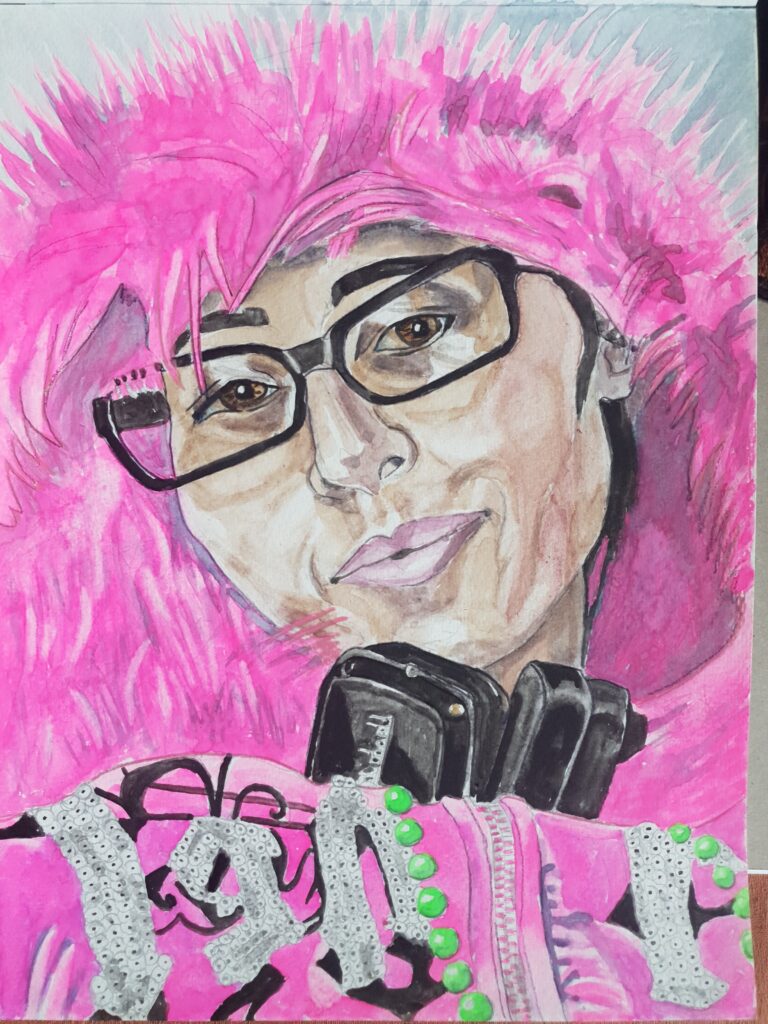 ID Woman wearing black glasses, she has a Mona Lisa smile, she is wearing a very fluffy furry fluorescent pink hoodie. In front of her is a fluorescent pink fabric with a zip in it which has sequinned shapes on it and some green beads hanging down it.