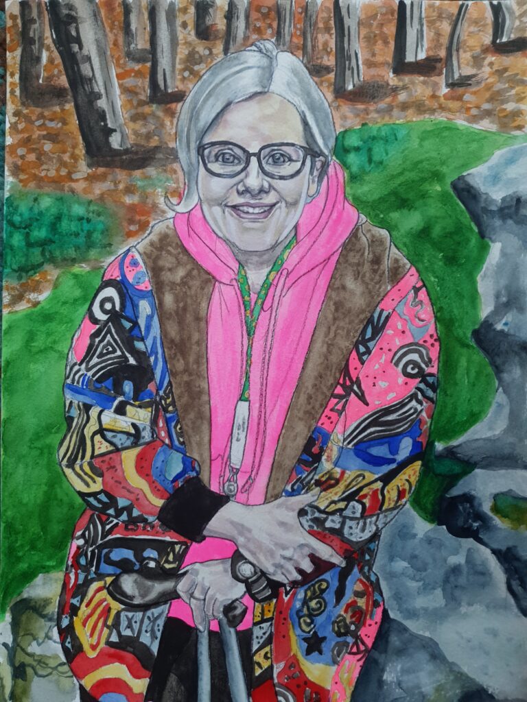 Woman with short blonde bob, she is smiling and is wearing glasses and a very colourful coat, she is out in the countryside with trees and leaning on a rock - which is on grass and ther is foliage and trees in the background