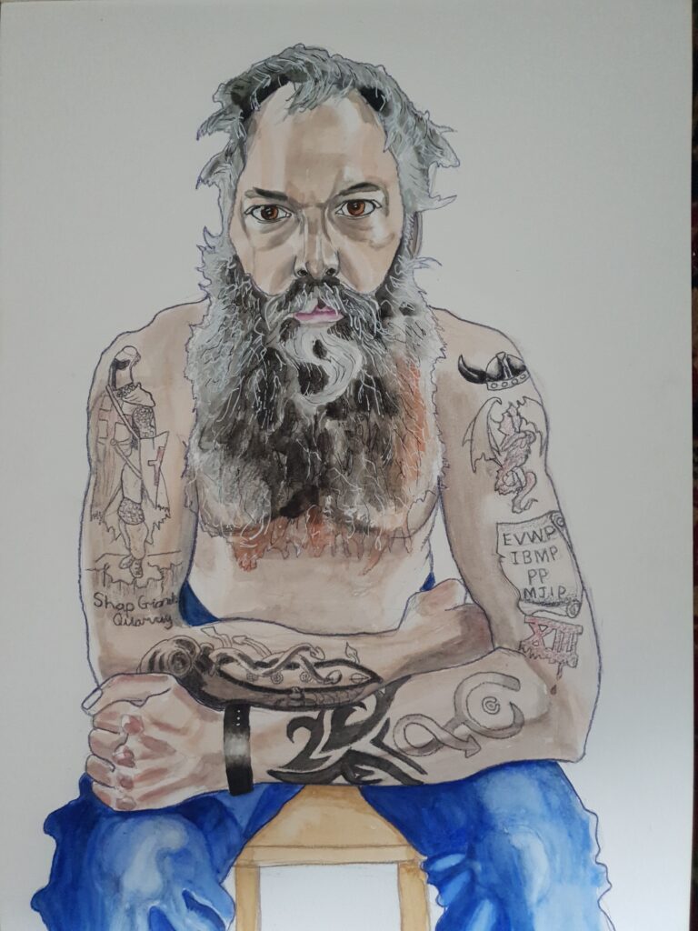 ID Portrait of a man sat on a stool - he is leaning forward with his arms bent resting on his legs. His arms are covered in tatoos. He has a very long full beard and scraggy hair, both are grey, he is wearing blue jeans and no top .