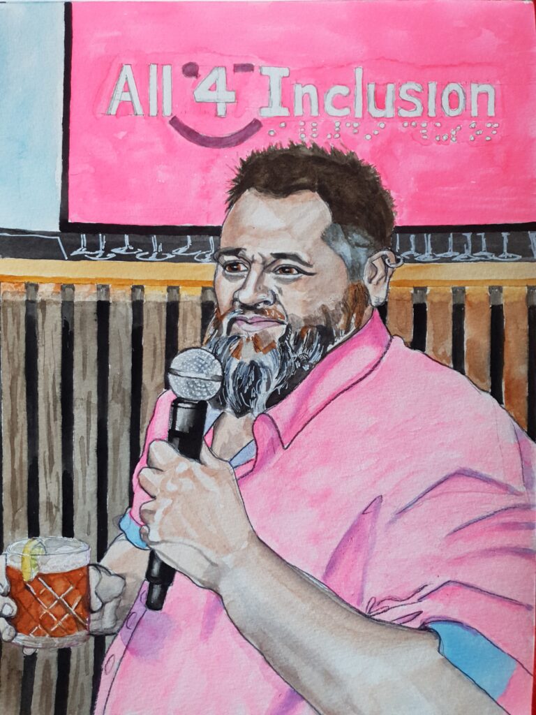 ID Well build man with short spiky hair and multicoloured beard. He is viewed at 3/4 angle and is sat down. He is holding a tumbler in one hand with some orange/brown liquid in it and a slice of lemon on the side. In his other hand he is holding a microphone. He is wearin ga pink shirt and behind him is a bar where you can see glasses showing beneath a sign which reads All 4 Inclusion, which is the organisation that Scott set up, to help disabled people experiencing loneliness.