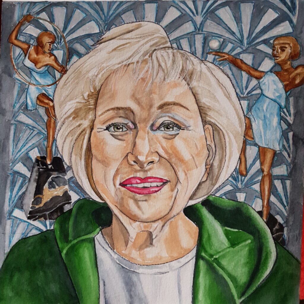 ID Mature woman with bouffant blonde bob, she is wearing a green hoodie, with grey t-shirt underneath - she is smiling nad is wearing red/pink lipstick. In the background can be seen 2 antique 20s figurines and a 20s style wallpaper