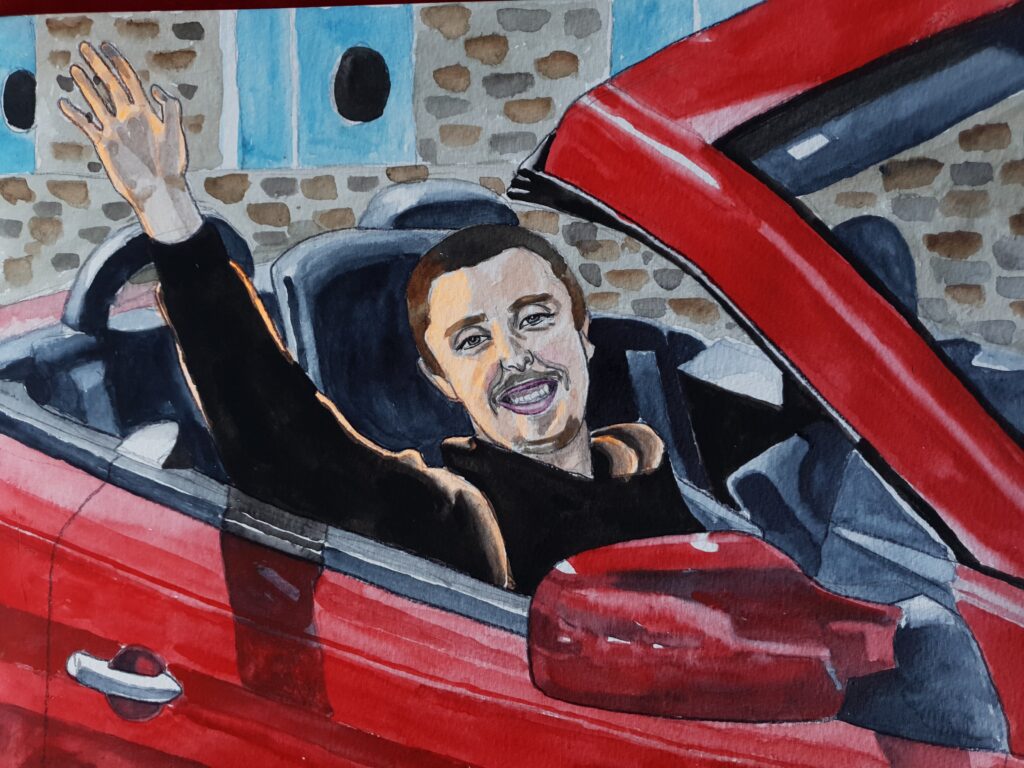 ID Man with short dark brown hair and a moustache smiling and waving from a red car the car is cropped and it is one with the top down. In the background can be seen rustic cottages with light blue doors. The car shapes make an excellent frame for the portrait.