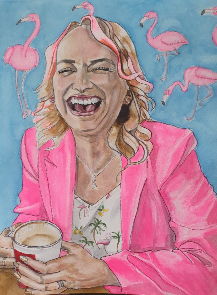 ID Young woman laughing her head off with a very infections expression, she has long wavy blonde hair with hints of pink and is wearing a bright pink jacket. Underneath her jacket is a white v necked top with flamingos and palm trees on it. She is holding a mug of coffee. In the background are flamingos in various stances which are pink on a blue background.